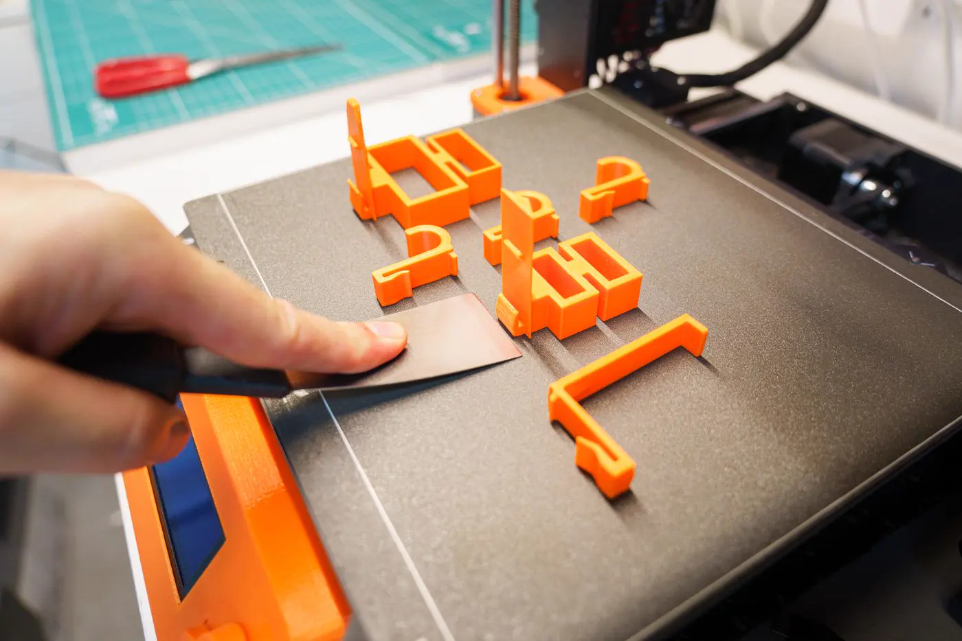 Une main décolle à la spatule des pièces orange du plateau d'une imprimante 3D