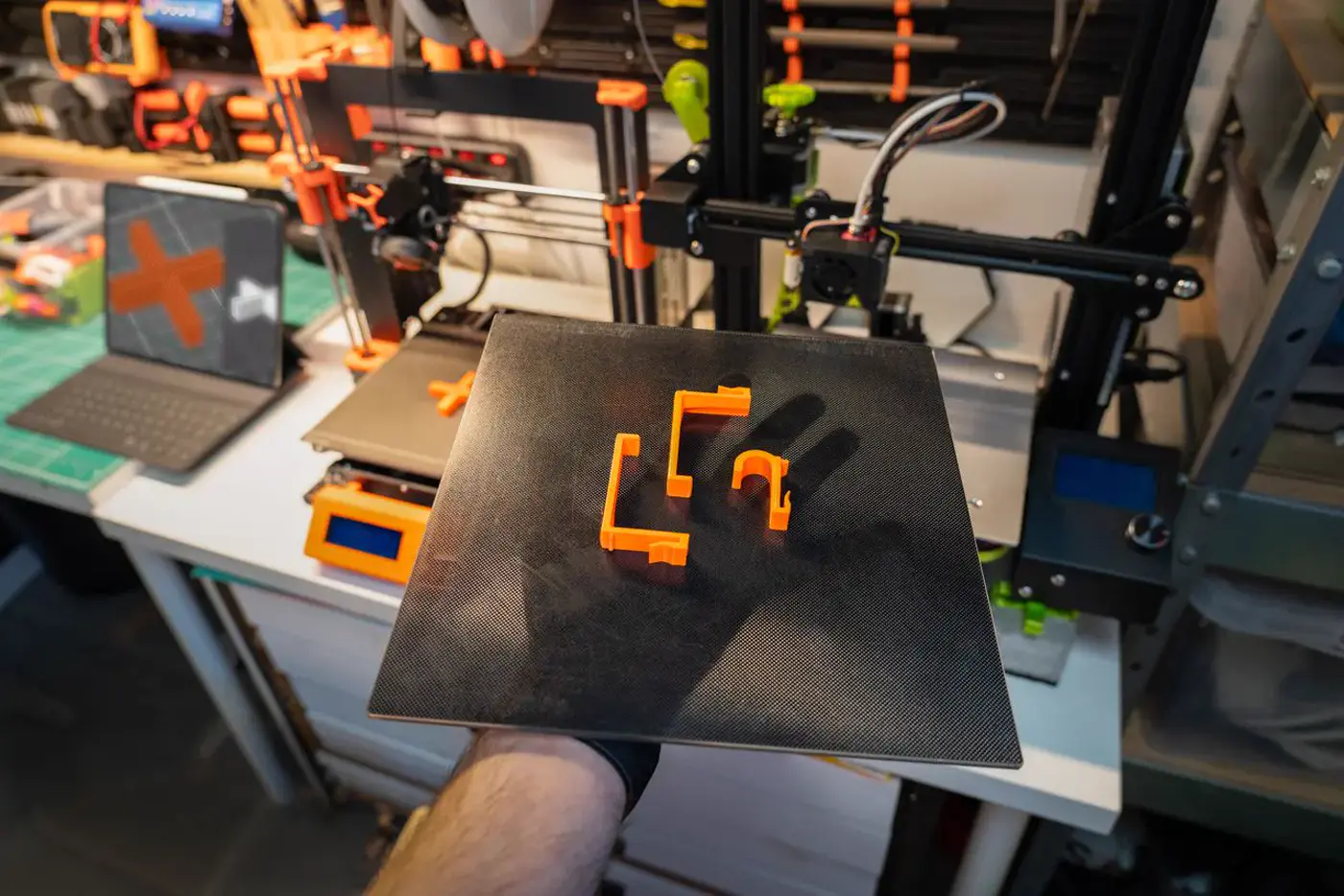 Une main gantée tient un plateau d'impression noir portant de petites pièces orange, devant une imprimante 3D
